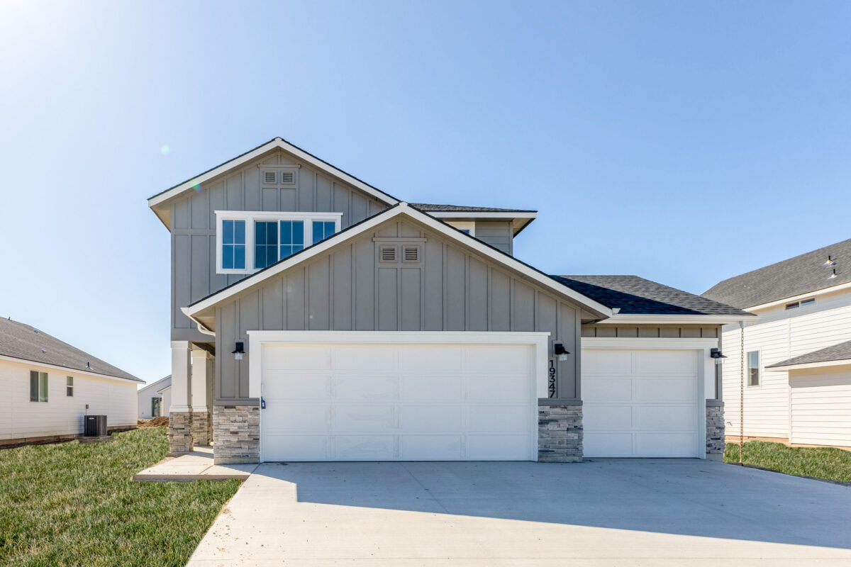 Modern two-story home with a three-car garage in a newly built community. The house features a gray and white exterior with stone accents, large windows, and a well-maintained front yard under a clear blue sky.