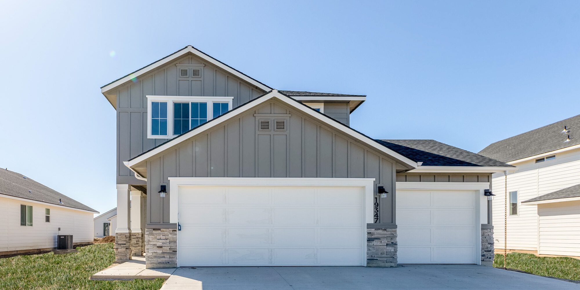 Modern two-story home with a three-car garage in a newly built community. The house features a gray and white exterior with stone accents, large windows, and a well-maintained front yard under a clear blue sky.