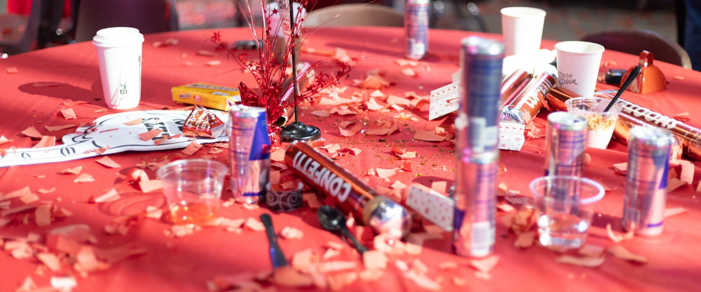 A close-up of a table at the 2025 CBH Homes Company Kickoff event, covered in red decorations, confetti, and party poppers. Empty energy drink cans, disposable coffee cups, snack wrappers, and celebration remnants create a lively post-event atmosphere. The scene captures the excitement and high energy of the CBH Homes corporate gathering, emphasizing a culture of enthusiasm, teamwork, and motivation within the homebuilding industry.
