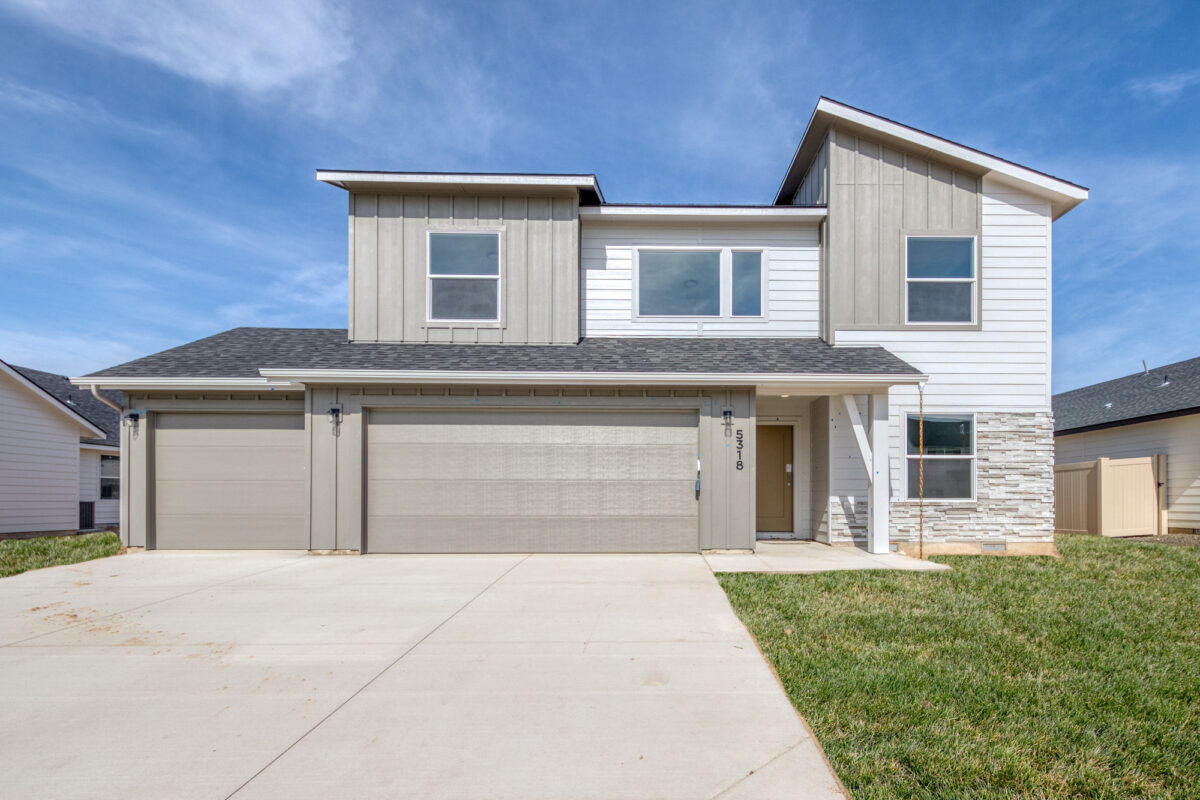 Modern two-story home with a three-car garage, light gray and white exterior, and stone accents. The house features a covered front porch, large windows, and a well-maintained lawn under a bright blue sky.